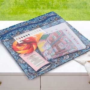 May include: A blue and floral zippered pouch with a clear plastic window. The pouch contains a variety of craft supplies, including embroidery floss, needles, and a cross-stitch pattern.