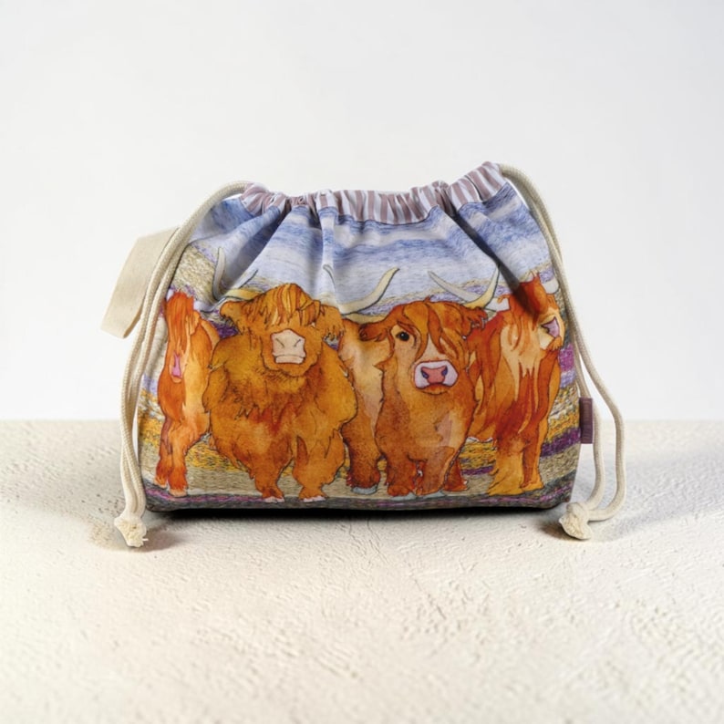 Op de afbeelding: Een stoffen buidel met trekkoord voorzien van een aquarelillustratie van vier bruine Schotse hooglanders op een rij. De tas heeft een geplooide bovenkant met cr&egrave;mekleurige katoenen koorden en een lichtblauwe en witte lucht.