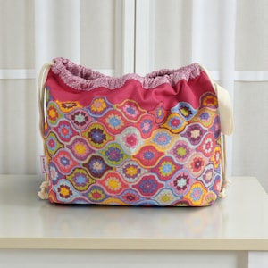 May include: A drawstring bag featuring a vibrant, multi-coloured crochet-style pattern of interlocking scalloped shapes in shades of pink, yellow, blue, and purple. The bag has a solid dark pink top section and thick, cream-coloured rope drawstrings.
