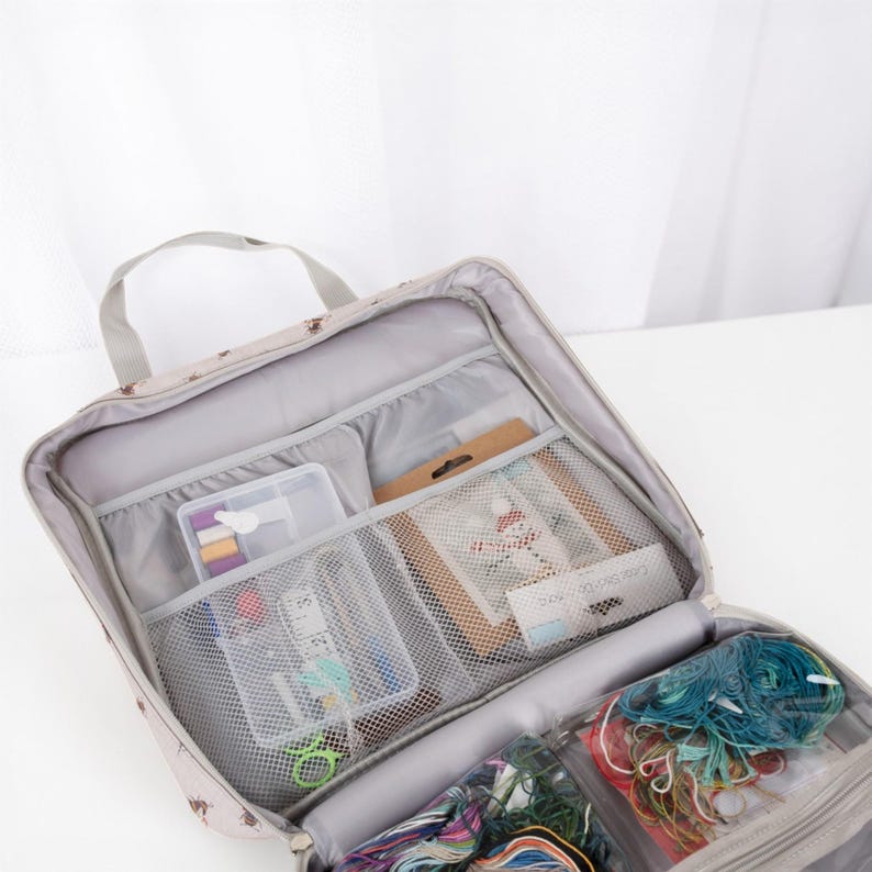 May include: Beige craft organiser bag with multiple compartments. The bag has a small pattern and a grey handle. Inside are various craft supplies, including thread, scissors, and small containers. The bag is open, displaying the contents.