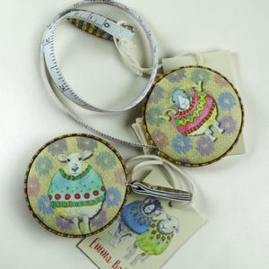 May include: Two round retractable measuring tapes featuring illustrations of sheep wearing colourful sweaters. One tape is extended, showing markings in cm. Each tape is attached to a small card with the text Emma Ball and sheep graphics.