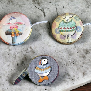 May include: Three colorful metal tape measures with different designs. One features a person in a red and white outfit sitting on a mushroom, another features a white sheep wearing a green sweater, and the last one features a puffin wearing a blue and white sweater.