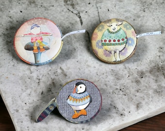 エマ・ボール作「TAPE MEASURES」。ノーム、セーターを着た羊、ミトンをはいた子猫、そしてウールのツノメドリのデザインもご用意しています。