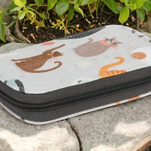 May include: A gray zippered pouch with a white background and a repeating pattern of cats and yarn balls. The pouch is lying on a gray stone surface.