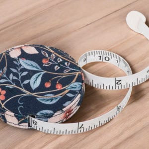 Può includere: Un metro a nastro rotondo con motivo floreale con un metro a nastro bianco esteso. Il metro a nastro è contrassegnato in centimetri, che mostra fino a 25 cm. L'involucro del nastro è blu navy con un design floreale in rosa, pesca e verde acqua.