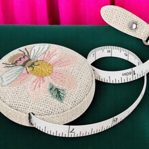 Op de afbeelding: Een ronde, beige meetlint met een bloemen- en bijenborduurontwerp. De witte tape loopt tot 152 cm. De tape-etui heeft een bijpassende beige band met een metalen druksluiting. De achtergrond is groen en roze.