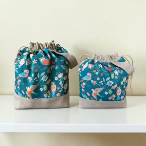 May include: Two drawstring bags in different sizes, featuring a teal fabric with a repeating pattern of colourful birds, flowers, and leaves. Each bag has a solid beige fabric base and a matching beige drawstring closure at the top opening.