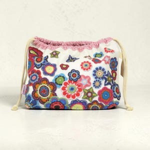 May include: A white drawstring pouch featuring a vibrant, multi-colored floral pattern with various crocheted-style flower motifs in shades of red, blue, green, and pink. The bag has a pink crocheted top edge and cream-colored cord drawstrings.