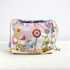 May include: A fabric drawstring pouch featuring a whimsical garden scene with multiple gnomes, large colorful flowers, and a watering can. The bag has a light purple patterned top border, cream-colored drawstrings, and a small purple side tag.