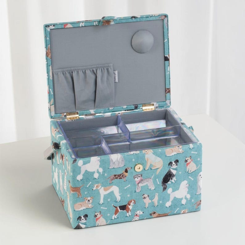 May include: A teal fabric storage box featuring a repeating pattern of various dog breeds. The box has a hinged lid with a gray interior, a small pocket, and a clear plastic removable tray with multiple compartments for organizing small items.