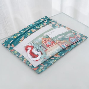 May include: A teal cross-stitch kit with a floral pattern and a zip closure. The kit includes a partially completed cross-stitch project depicting beach houses, along with white fabric, embroidery floss, and a pattern guide. The kit measures approximately 28 x 28 cm.
