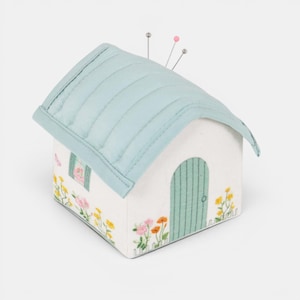 May include: A house-shaped pin cushion made of white fabric, featuring a light blue quilted roof and a green door. The house is adorned with embroidered flowers and has pins inserted into the roof.