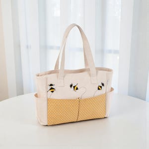 Può includere: Borsa tote beige con api ricamate. La borsa presenta una tasca a pois gialli e due tasche laterali. I manici e il corpo principale della borsa sono beige. Le api sono nere e gialle.