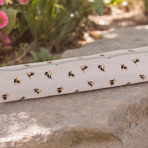 May include: A gray linen zippered pouch with a repeating pattern of black and yellow bumblebees. The pouch is lying on a gray rock.
