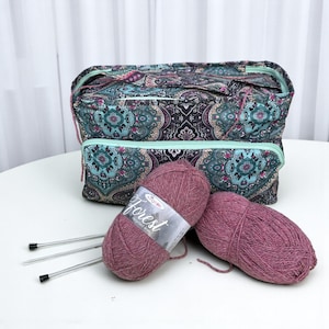 Puede incluir: Una bolsa de tejido de tela estampada con cremalleras turquesas y dos madejas de hilo malva. El hilo está etiquetado como "Forest". También hay tres agujas de tejer plateadas.