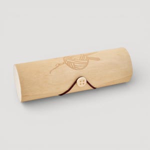 Könnte beinhalten: Ein helles Stricknadel-Etui aus Holz mit einem lasergravierten Design aus Garn und Nadeln. Das Etui ist zylindrisch mit einem Knopfverschluss und einer dunkelbraunen Kordel. Das Etui ist aus Holz und hat eine natürliche Oberfläche.