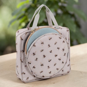 May include: A light grey craft bag with a bee pattern, holding a round embroidery hoop. The bag has a handle and a zipped compartment. The embroidery hoop is light blue and wooden.