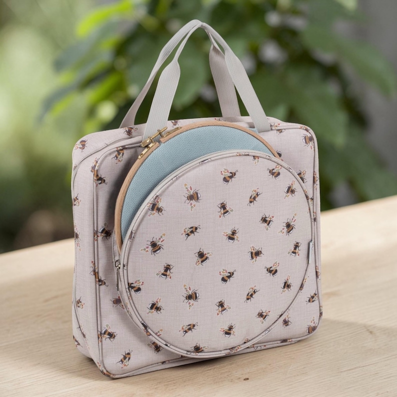 May include: A light grey craft bag with a bee pattern, holding a round embroidery hoop. The bag has a handle and a zipped compartment. The embroidery hoop is light blue and wooden.