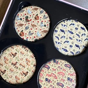 Peut inclure: Ensemble de quatre sous-verres ronds avec différents motifs d'animaux de ferme. Les sous-verres présentent un fond beige avec des illustrations colorées de poulets, de vaches, de moutons et de cochons.