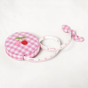 May include: A round retractable measuring tape covered in pink and white gingham fabric with a small embroidered cherry motif on top. The white measuring tape extends from the case, showing markings in centimetres up to 150 cm in total length.