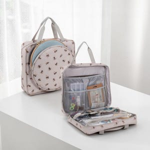 May include: Two beige craft bags with a bee pattern. One bag has a circular opening for an embroidery hoop. The other bag is open, revealing pockets and compartments for storing craft supplies. Both bags have grey handles.