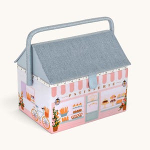 Könnte beinhalten: Ein rechteckiger Nähkorb mit hausförmigem Deckel und blauem Griff. Die Außenseite zeigt eine Illustration einer rosa Patisserie mit Fahrrad, Gebäck und dem Schriftzug PATISSERIE. Ein Maßband ist entlang der Deckelkante aufgedruckt.