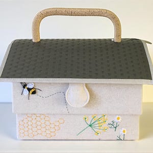 May include: A fabric box with a woven handle, featuring a gray roof-like top and a cream-colored body. The box is decorated with embroidered bees, a honeycomb, and floral designs. The handle is made of a light brown woven material.