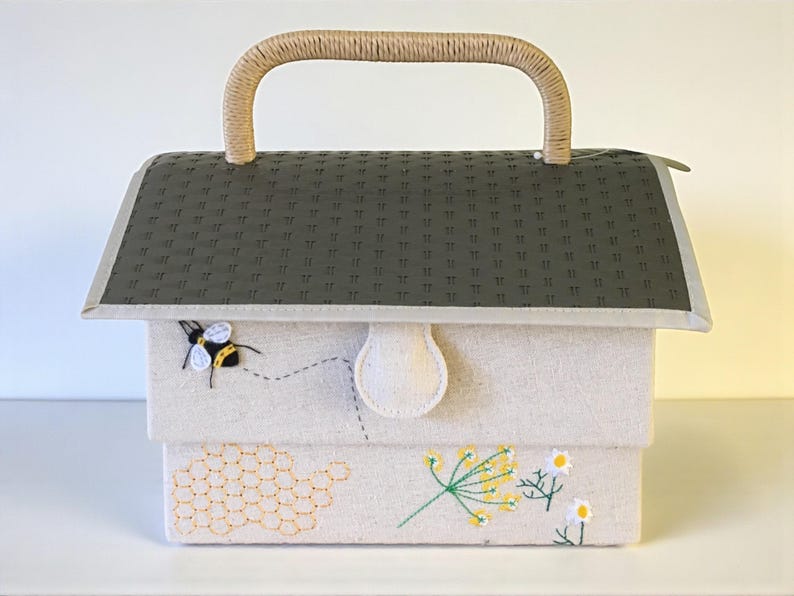 May include: A fabric box with a woven handle, featuring a gray roof-like top and a cream-colored body. The box is decorated with embroidered bees, a honeycomb, and floral designs. The handle is made of a light brown woven material.