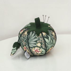 May include: A round tomato-shaped pincushion featuring a dark green fabric with a floral and leaf pattern in shades of yellow, pink, and light green. It has a dark green felt top, a small matching leaf attachment, and three silver sewing pins.