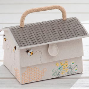 May include: A beige fabric house-shaped box with a woven handle. The roof is gray with a textured pattern. The box is decorated with embroidered bees, honeycomb, and flowers. The box has a clasp closure.