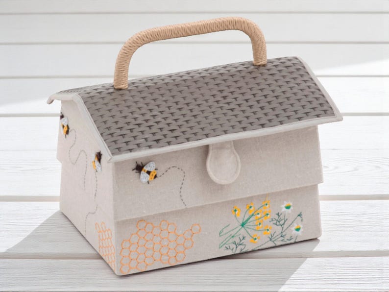 May include: A beige fabric house-shaped box with a woven handle. The roof is gray with a textured pattern. The box is decorated with embroidered bees, honeycomb, and flowers. The box has a clasp closure.