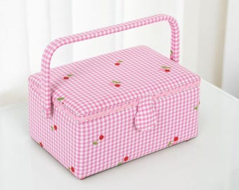 May include: A pink gingham sewing box with a handle and cherry embroidery. The box has a rectangular shape with a curved handle and a button closure. The cherries are red with green stems.