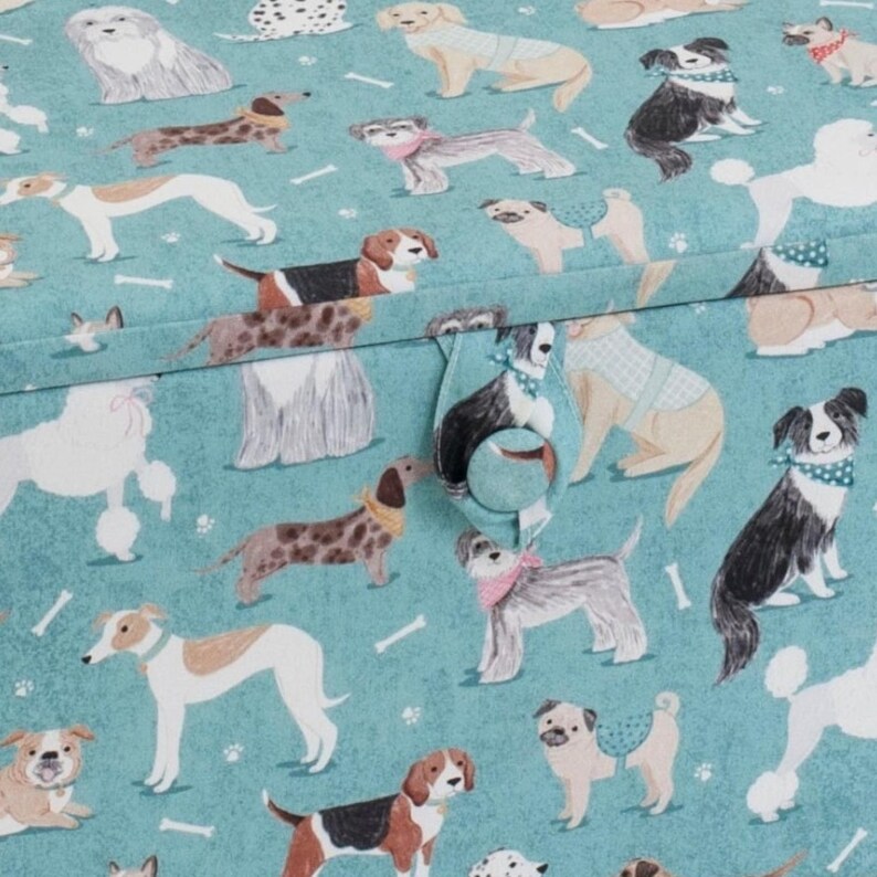 May include: A teal fabric featuring a repeating pattern of various dog breeds, including poodles, pugs, and beagles, interspersed with small white bone shapes. A fabric tab with a matching circular button is centered on the front surface.