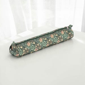 Könnte beinhalten: Ein langes, rechteckiges Federmäppchen mit einem Blumenmuster in Grün-, Rosa- und Cremetönen. Das Etui hat einen dunkelgrünen Reißverschluss und Besatz. Das Federmäppchen ist aus Stoff gefertigt und für Stifte, Kugelschreiber und andere Schreibwaren konzipiert.