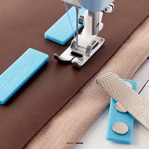 Pu&ograve; includere: Una macchina da cucire con ago e piedino premistoffa &egrave; posizionata su tessuto marrone e una cerniera beige. Sono visibili anche due guide magnetiche per cuciture Hemline blu e una guida magnetica per cuciture argentata.