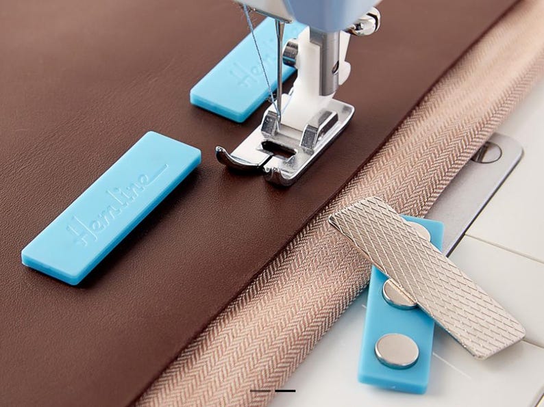 Pu&ograve; includere: Una macchina da cucire con ago e piedino premistoffa &egrave; posizionata su tessuto marrone e una cerniera beige. Sono visibili anche due guide magnetiche per cuciture Hemline blu e una guida magnetica per cuciture argentata.