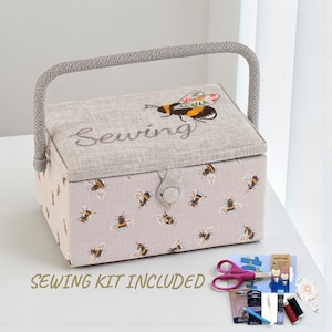 Peut inclure: Une boîte à couture beige avec une poignée grise et un couvercle brodé d'une abeille et du mot "Sewing". La boîte est ornée de motifs d'abeilles. Un kit de couture est inclus.