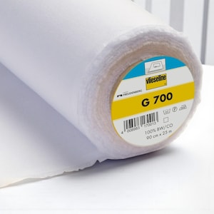 Könnte beinhalten: Eine Rolle weiße Vlieseline G 700 Einlagestoff. Das runde Etikett am Ende zeigt den Markennamen, den Text G 700, einen Strichcode, die Spezifikationen 100% BW/CO, 90 cm x 25 m sowie verschiedene Pflegesymbole für die Textilreinigung.