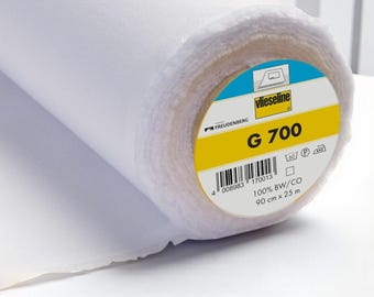 Peut inclure: Un rouleau de tissu thermocollant blanc Vlieseline G 700. L'étiquette circulaire à l'extrémité présente le nom de la marque, le texte G 700, un code-barres, les spécifications 100% BW/CO, 90 cm x 25 m, et des symboles d'entretien textile.