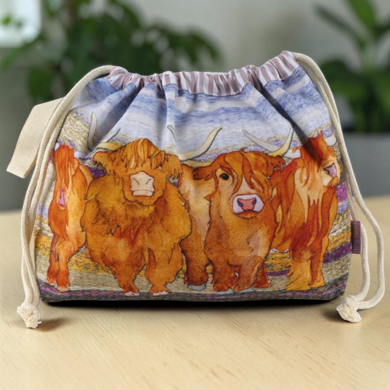 Op de afbeelding: Een stoffen buidel met trekkoord voorzien van een bedrukte illustratie van drie harige, gemberkleurige Schotse Hooglanders in een veld. De tas heeft een geplooide bovenkant met een lichtbruin en wit gestreept patroon en cr&egrave;mekleurige koorden.