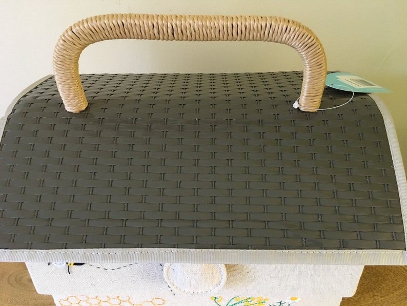 May include: A gray picnic basket with a woven pattern on the lid and a beige woven handle. The basket has a light-colored trim and a fabric base with embroidered bee and honeycomb designs. A tag hangs from the side.