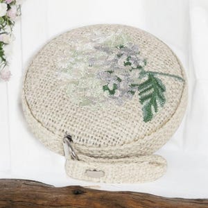 May include: A round, beige, woven bag with a floral embroidery design. The embroidery features a white flower with green leaves.