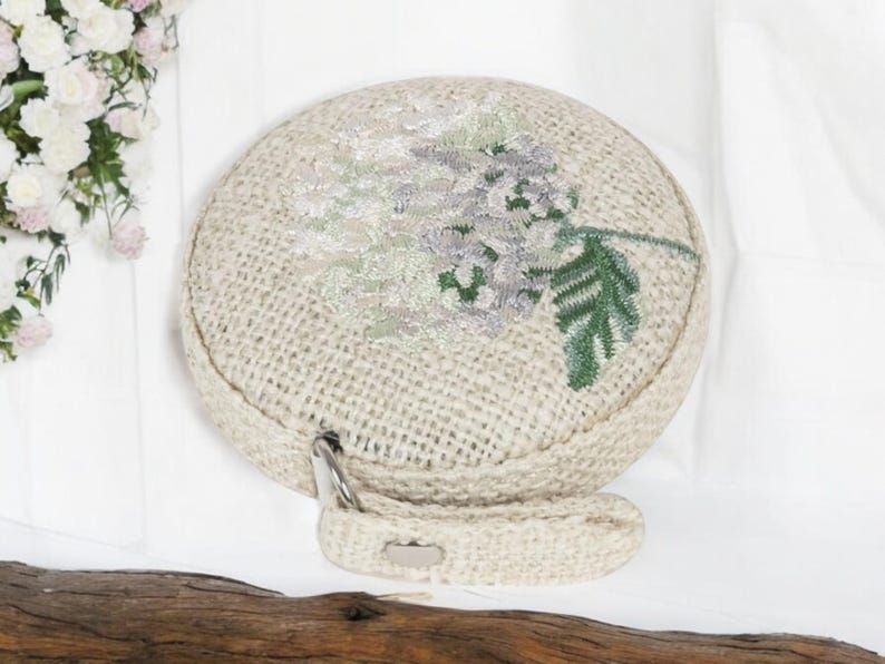 May include: A round, beige, woven bag with a floral embroidery design. The embroidery features a white flower with green leaves.