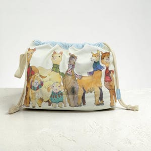 Könnte beinhalten: Ein cremefarbener Beutel mit Kordelzug, der mit einer Aquarellillustration von sechs Lamas und Alpakas in bunten Schals und Pullovern bedruckt ist. Die Tasche hat einen hellblauen Stoff mit Muster an der Öffnung und beigefarbene Kordeln.