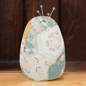 May include: A green and yellow patchwork pincushion with a floral and bee design. The pincushion has a round base and a pointed top with five silver pins.