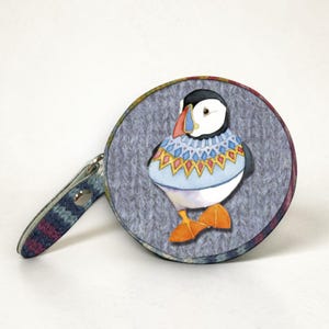 Peut inclure: Un objet rond, plat et circulaire présentant une illustration style aquarelle d'un macareux portant un pull à motifs bleu, jaune et rouge. Le fond de l'illustration est une texture tricotée grise. Une petite languette assortie est fixée.