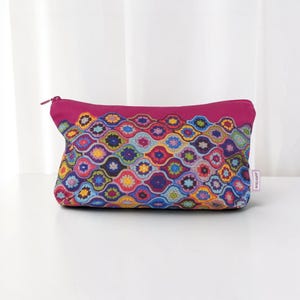 May include: A rectangular zippered pouch featuring a vibrant, multi-colored crochet-style pattern of interlocking scalloped shapes in shades of yellow, blue, red, and purple, with a solid magenta top border and a small white label on the side.