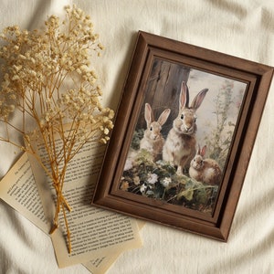 May include: A framed print of a painting depicting three rabbits in a woodland setting. The rabbits are brown and white with long ears and are surrounded by green foliage and white flowers.