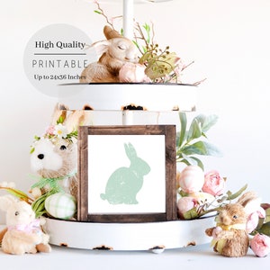Puede incluir: Una descarga digital imprimible de una silueta de conejo verde en un marco marr&oacute;n. El marco est&aacute; sentado en una bandeja de tres niveles blanca con decoraciones de Pascua, incluyendo figuritas de conejos y flores rosas.