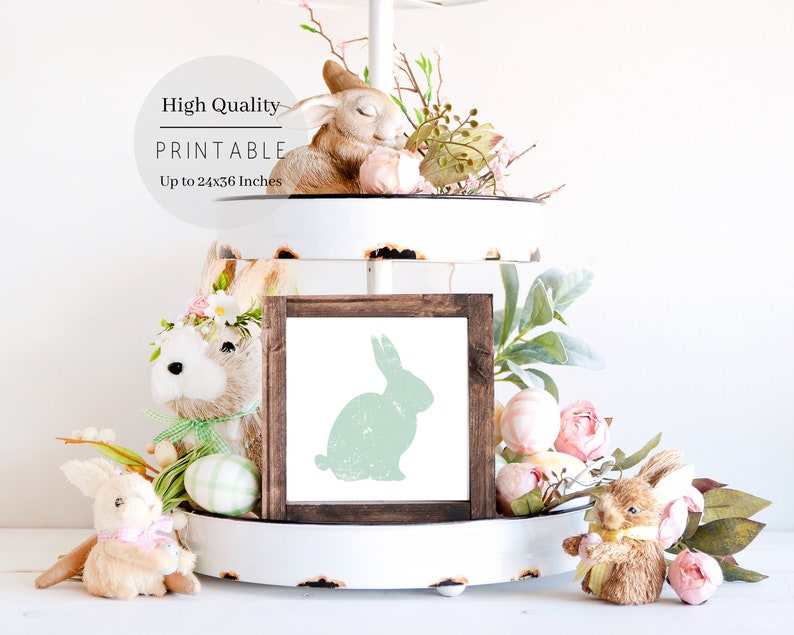 Puede incluir: Una descarga digital imprimible de una silueta de conejo verde en un marco marr&oacute;n. El marco est&aacute; sentado en una bandeja de tres niveles blanca con decoraciones de Pascua, incluyendo figuritas de conejos y flores rosas.
